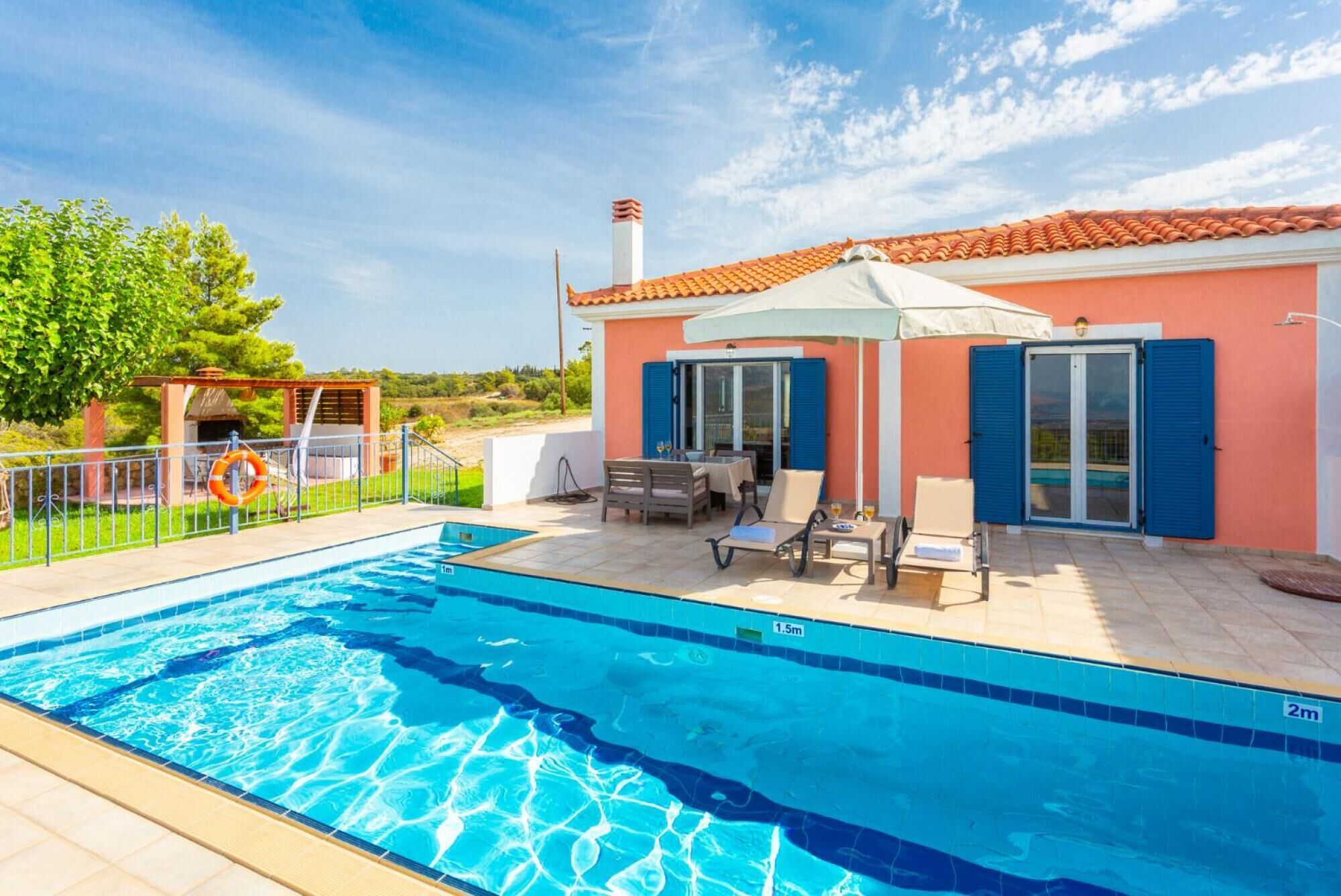 A swimming pool with a clear blue waterline, a covered patio area with outdoor furniture, and a building with a red-tiled roof and blue shutters.