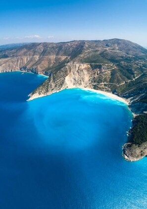 Vista aérea de uma paisagem costeira com mar azul límpido, montanhas ao fundo e uma praia estreita.