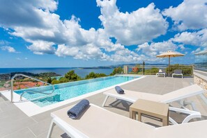 Piscina infinita no terraço com vista clara do mar e montanhas, cadeiras de espreguiçadeira e guarda-sol.