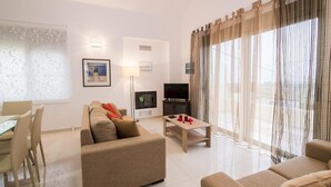 A modern living room with a sofa, a coffee table, a TV, and a dining area with chairs and a table. - Litsarda Villas in Apokoronos (Apokoronas)