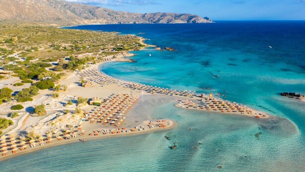 An aerial view of a coastal area with a beach, clear blue waters, and a mountainous backdrop. - Panormos Villas Collection (Mylopotamos)