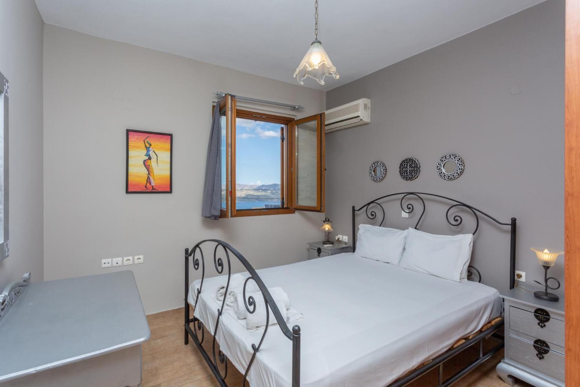 A bedroom with a bed, bedside table, lamp, air conditioning unit, and a window with a view.
