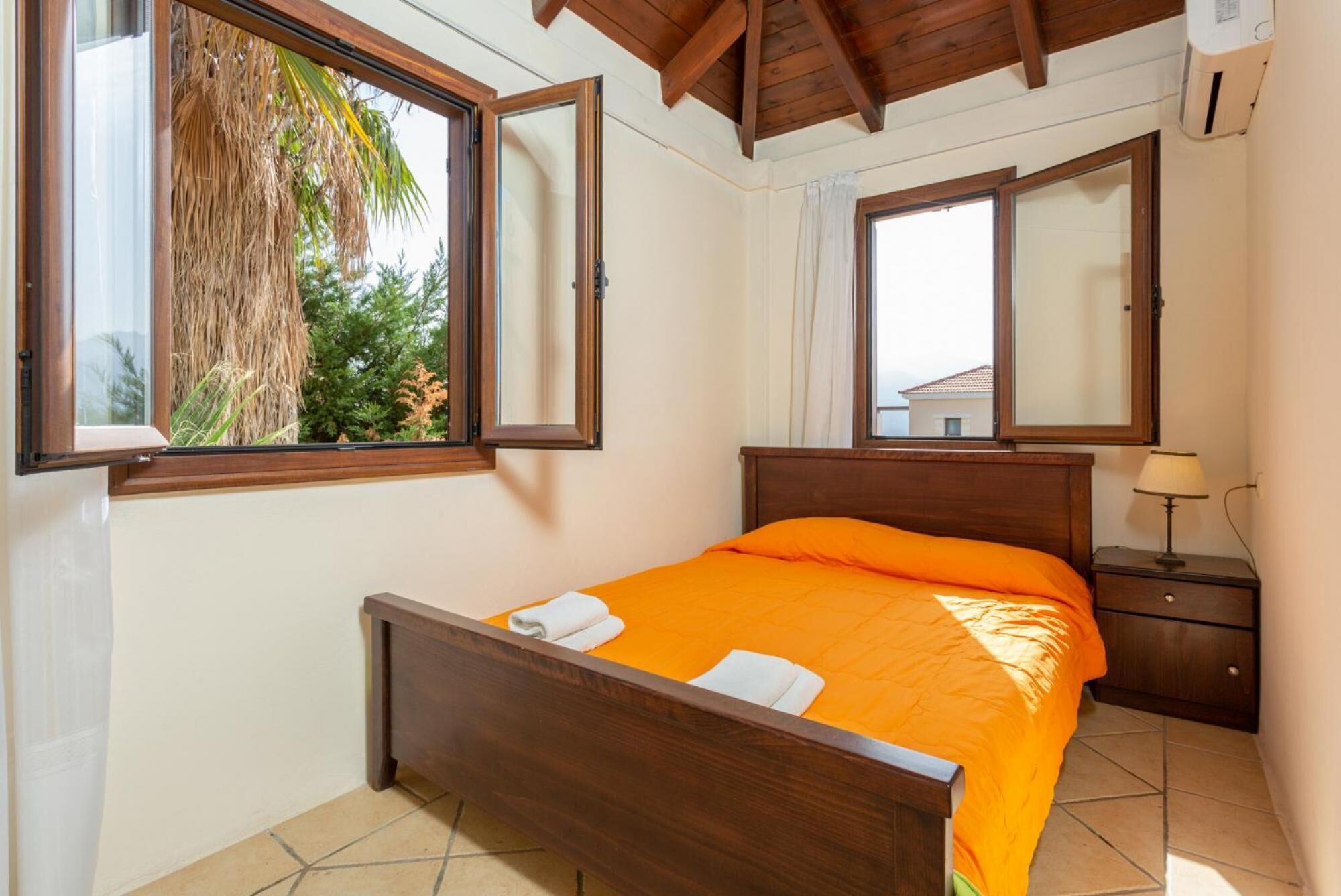 A bedroom with a wooden bed, a nightstand, a lamp, and a window with a view of palm trees.
