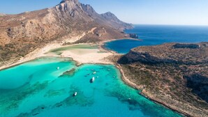 An aerial view of a coastal area with a prominent mountain, clear turquoise waters, and a sandy beach. - Spiros Villas Collection (Sfakia)