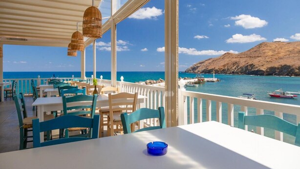 A seaside restaurant with outdoor seating, white tables, and blue chairs, overlooking the ocean and boats. - Spiros Villas Collection (Sfakia)
