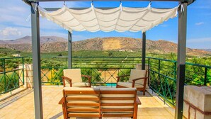 A terrace with two wooden chairs and a table, a pergola with a white canopy, and a view of green hills and mountains in the background. - Kalyves Villas Collection (Apokoronas)