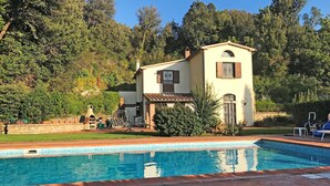 Blick vom Pool auf ein zweistöckiges Haus mit Poolfliesen, umgeben von Grünpflanzen und Bäumen.