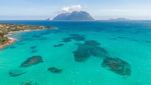 A coastal landscape with clear turquoise waters, rocky outcrops, and a prominent mountain in the distance.