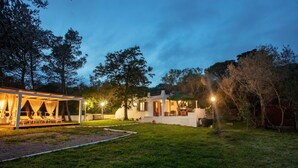 Un patio exterior moderno con mobiliario y una zona cubierta, rodeado de árboles y un césped cuidado.