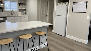 Una cocina moderna con encimera blanca, tres taburetes de madera, un refrigerador y un calendario en la pared.