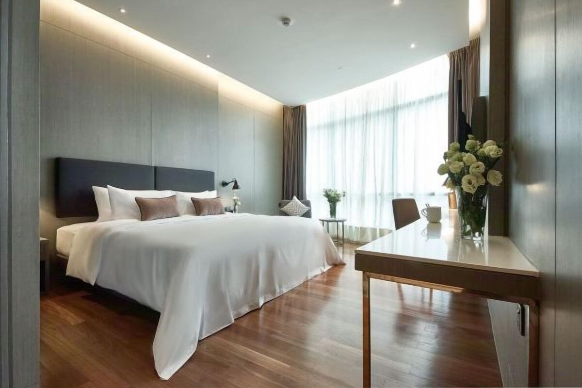 A modern hotel room with a large bed, a small table, and a view of the outdoors through large windows.
