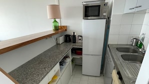 A compact kitchen with a microwave, refrigerator, sink, and a wooden shelf.