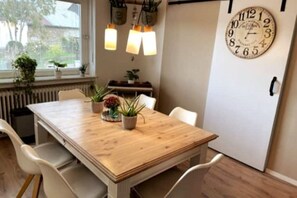 Ein Essbereich mit Holztisch, weißen Stühlen, Topfpflanzen, Pendelleuchten und einer Wanduhr.