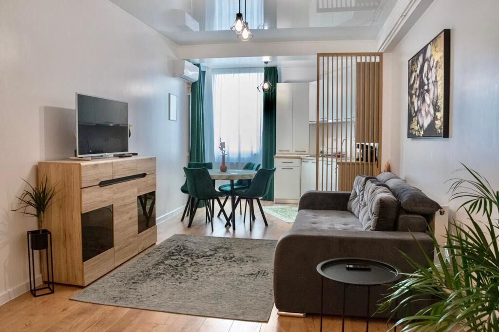 A modern living room with a sofa, a TV, a dining table with chairs, and a kitchen area visible through a sliding door.
