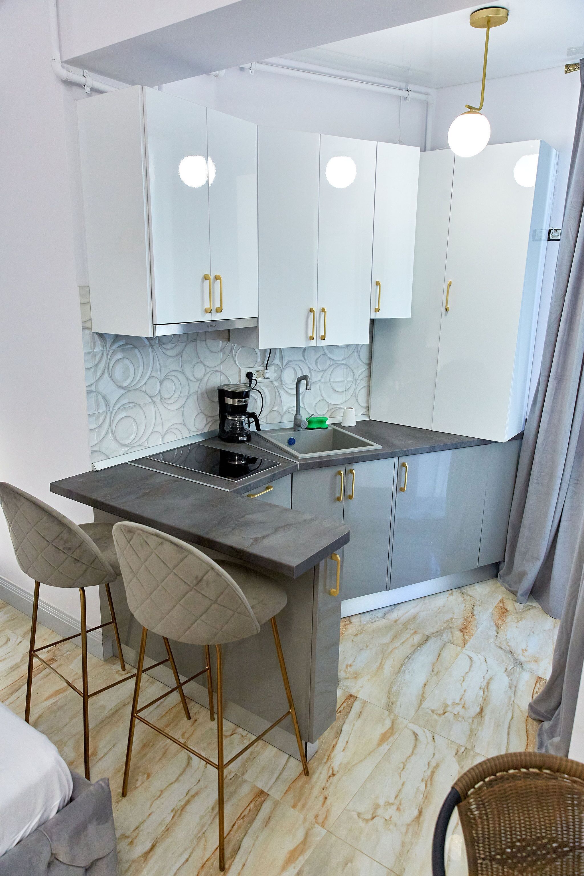A modern kitchen with white cabinets, a black countertop, and a coffee maker. There are two bar stools and a wicker chair.