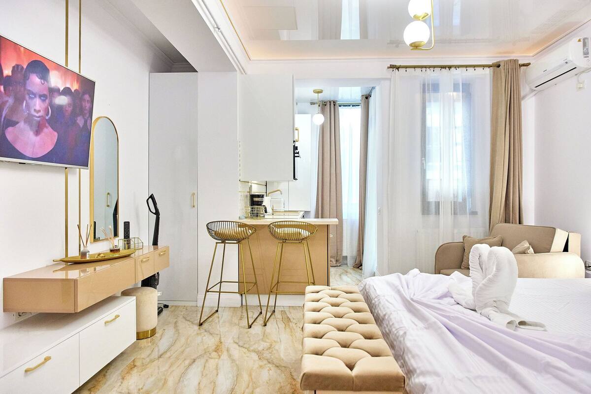 A modern hotel room with a large bed, a vanity with a mirror, a desk with two bar stools, and a television on the wall.