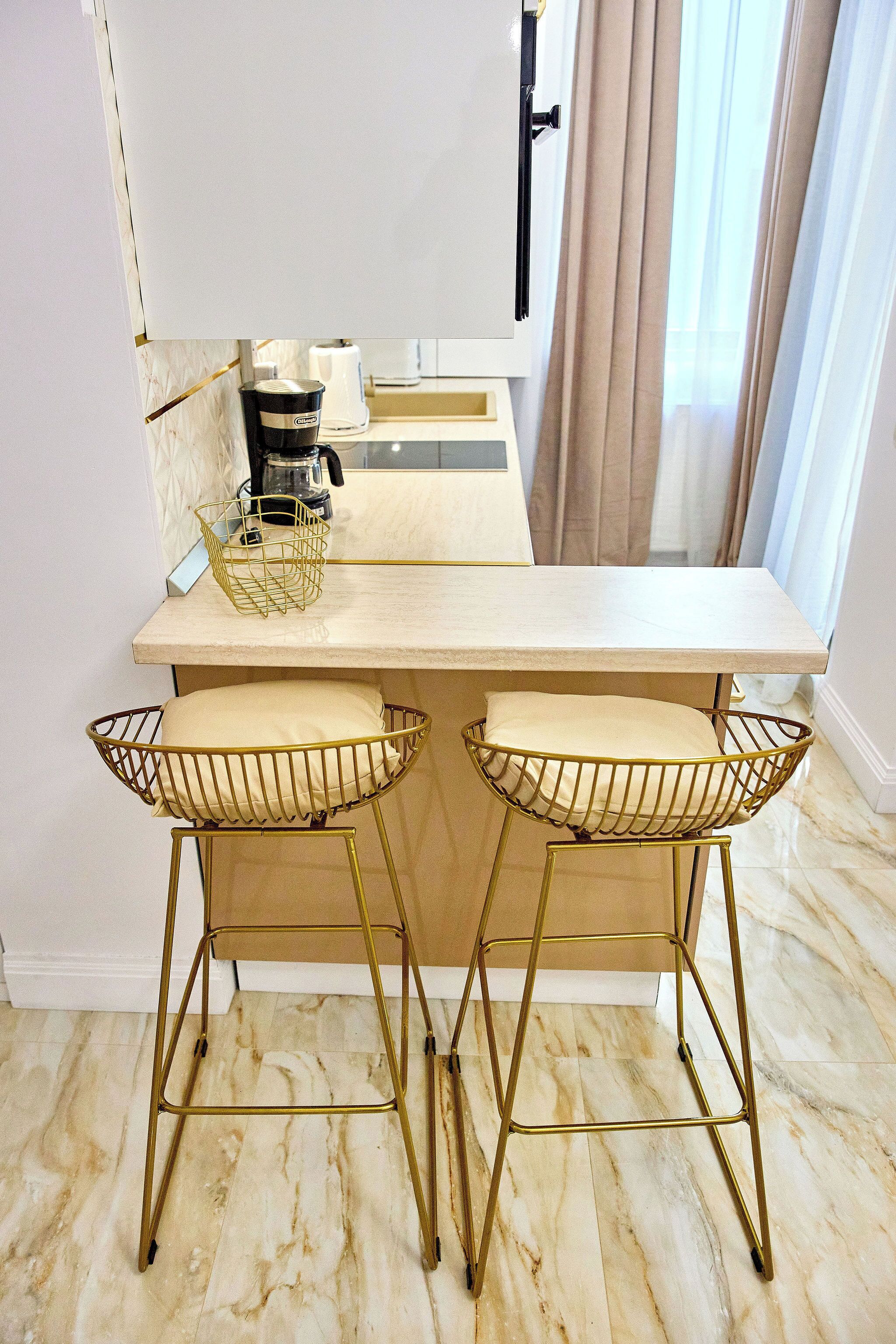 A kitchen area with a marble countertop, two bar stools with cushions, a coffee maker, and a basket on the counter.