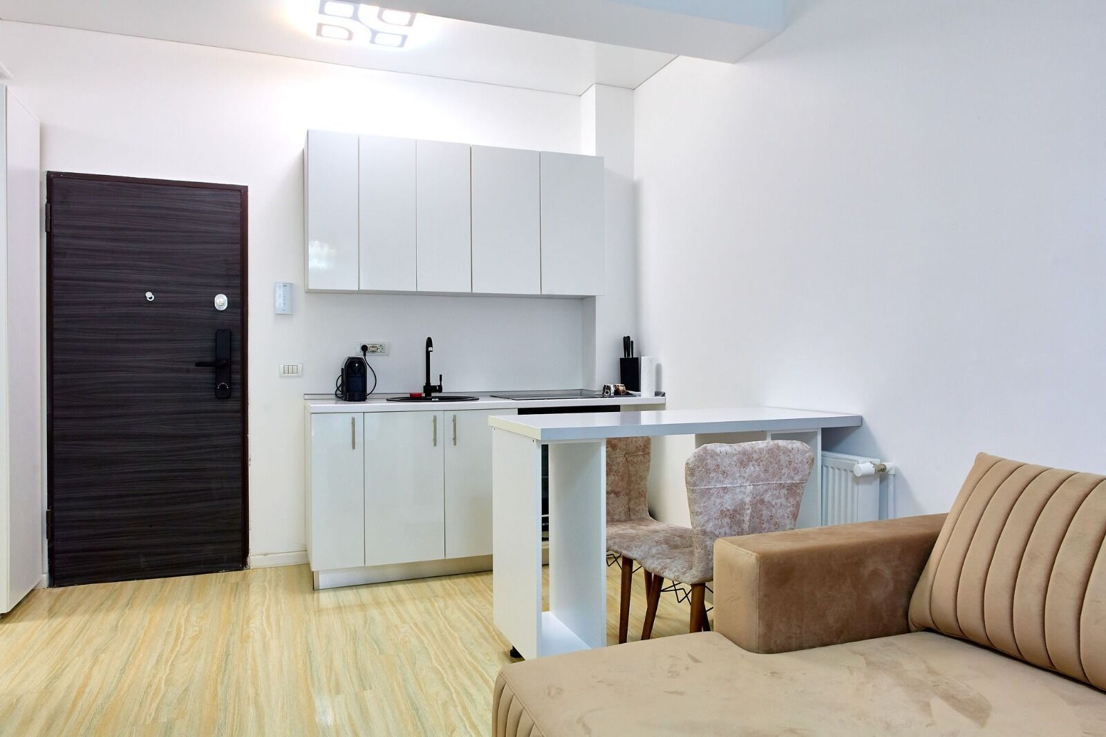 A compact living space with a kitchenette, a dark wooden door, a beige sofa, and a patterned chair.