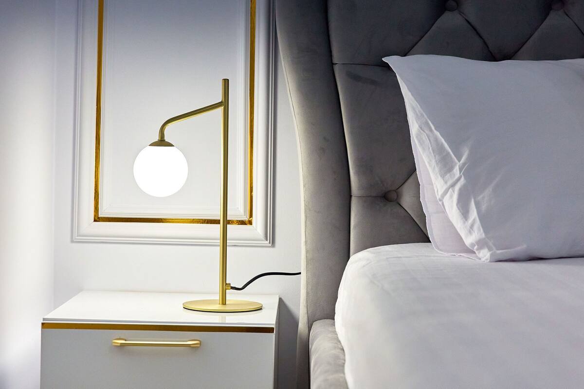 A neatly made bed with white bedding and a single white pillow, a bedside table with a gold lamp, and a framed picture on the wall.