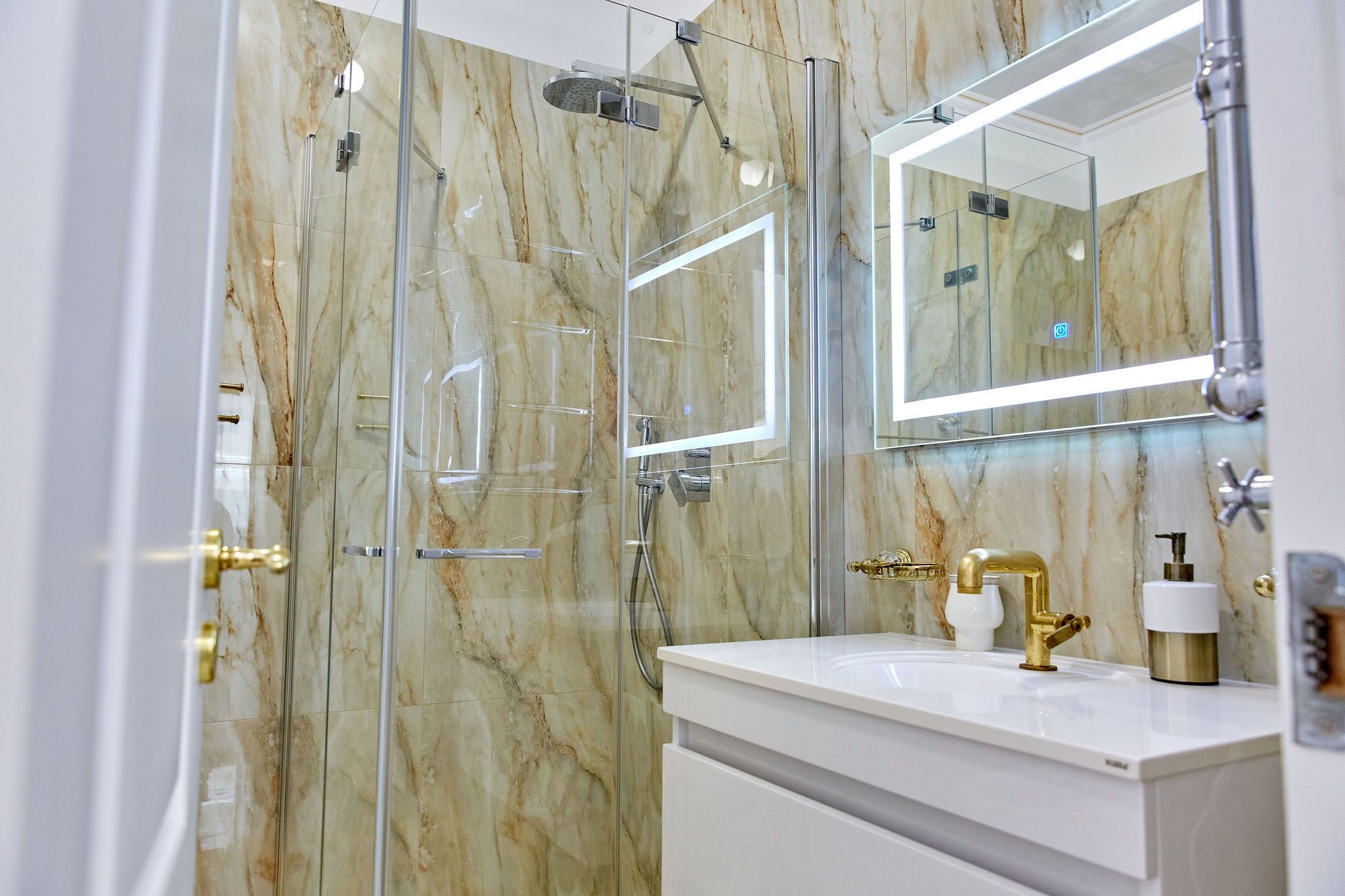 A modern bathroom with a glass-enclosed shower, marble walls, and a white vanity with gold fixtures.