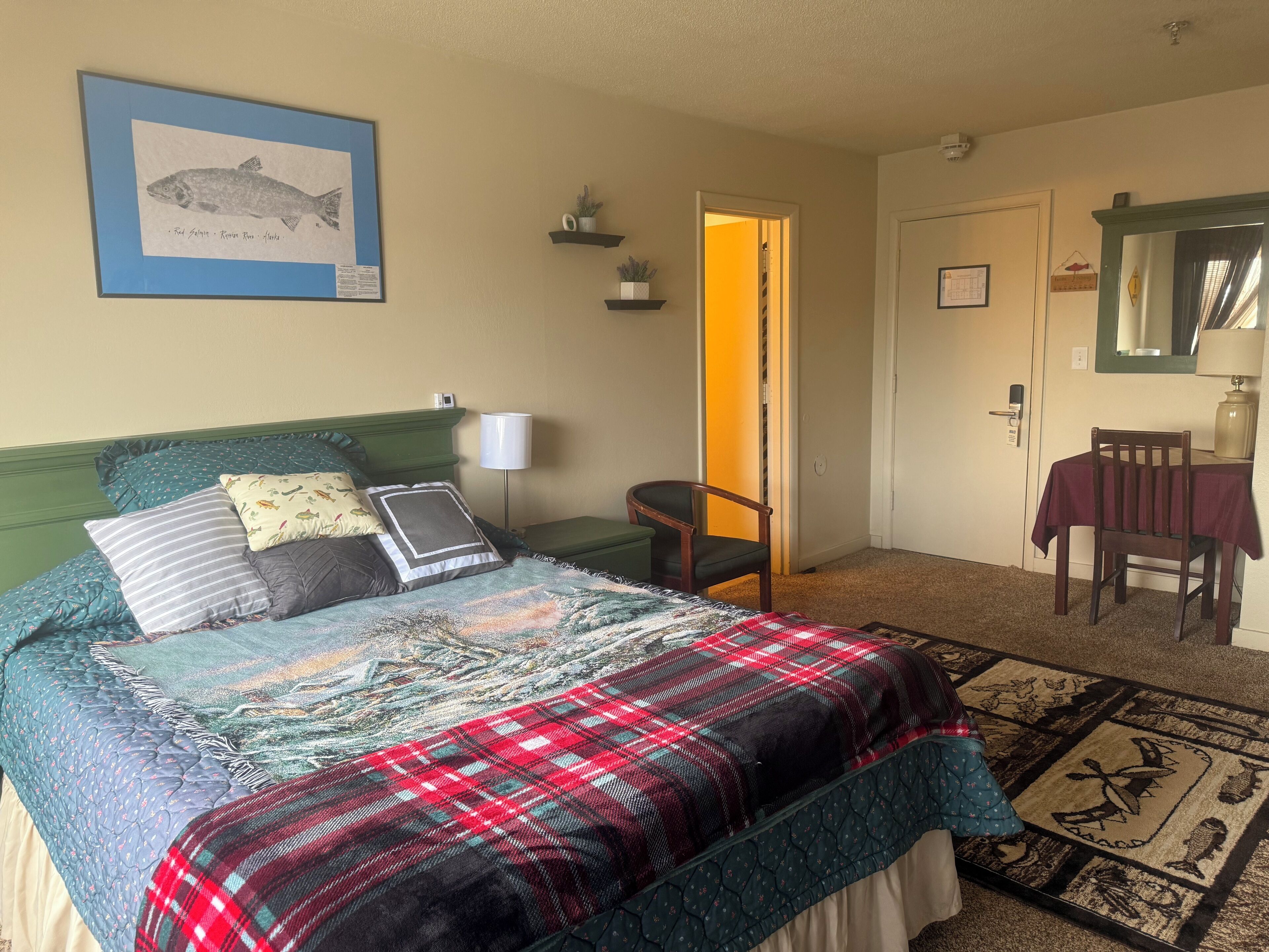 A bedroom with a bed, pillows, a bedside table, a chair, a desk with a tablecloth, a framed picture on the wall, and a carpet with a rug.