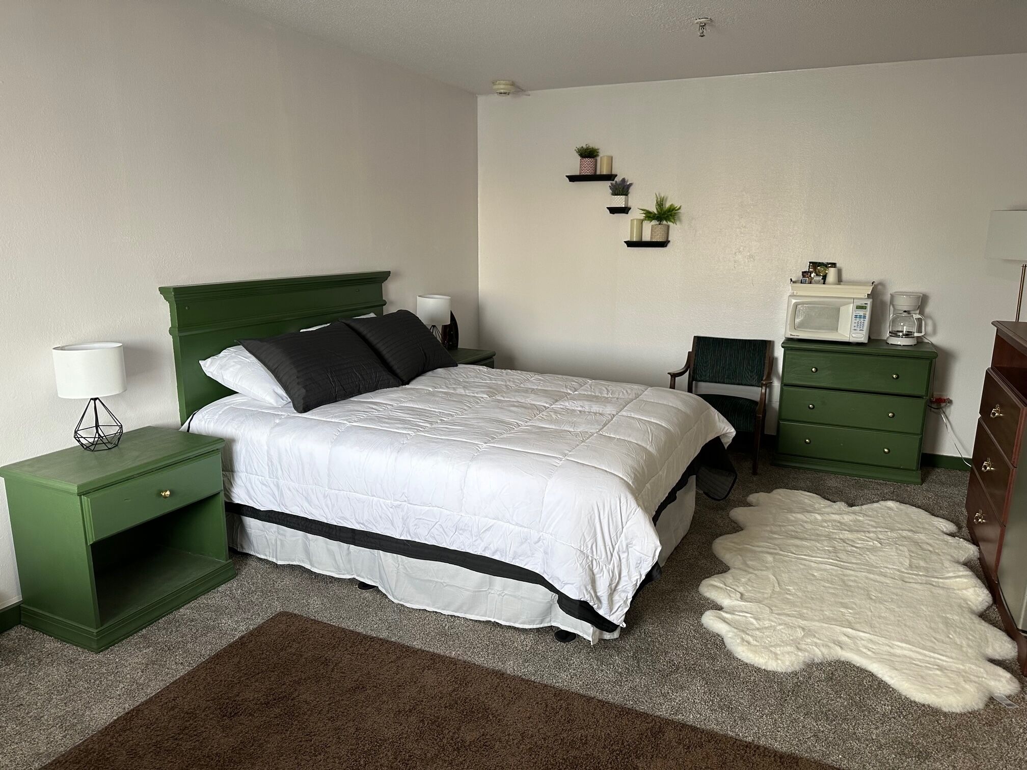 Une chambre à coucher bien rangée, avec un lit vert, une table de chevet avec une lampe, une commode, un four à micro-ondes et une petite étagère ornée d’objets décoratifs.