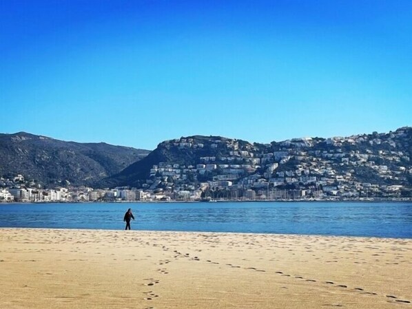A beach with footprints leading to the water, a person standing, and a hillside with buildings.