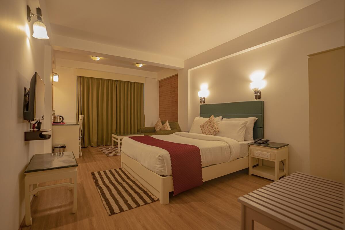 A hotel room with a bed, bedside table, nightstand, and a small desk with a chair.