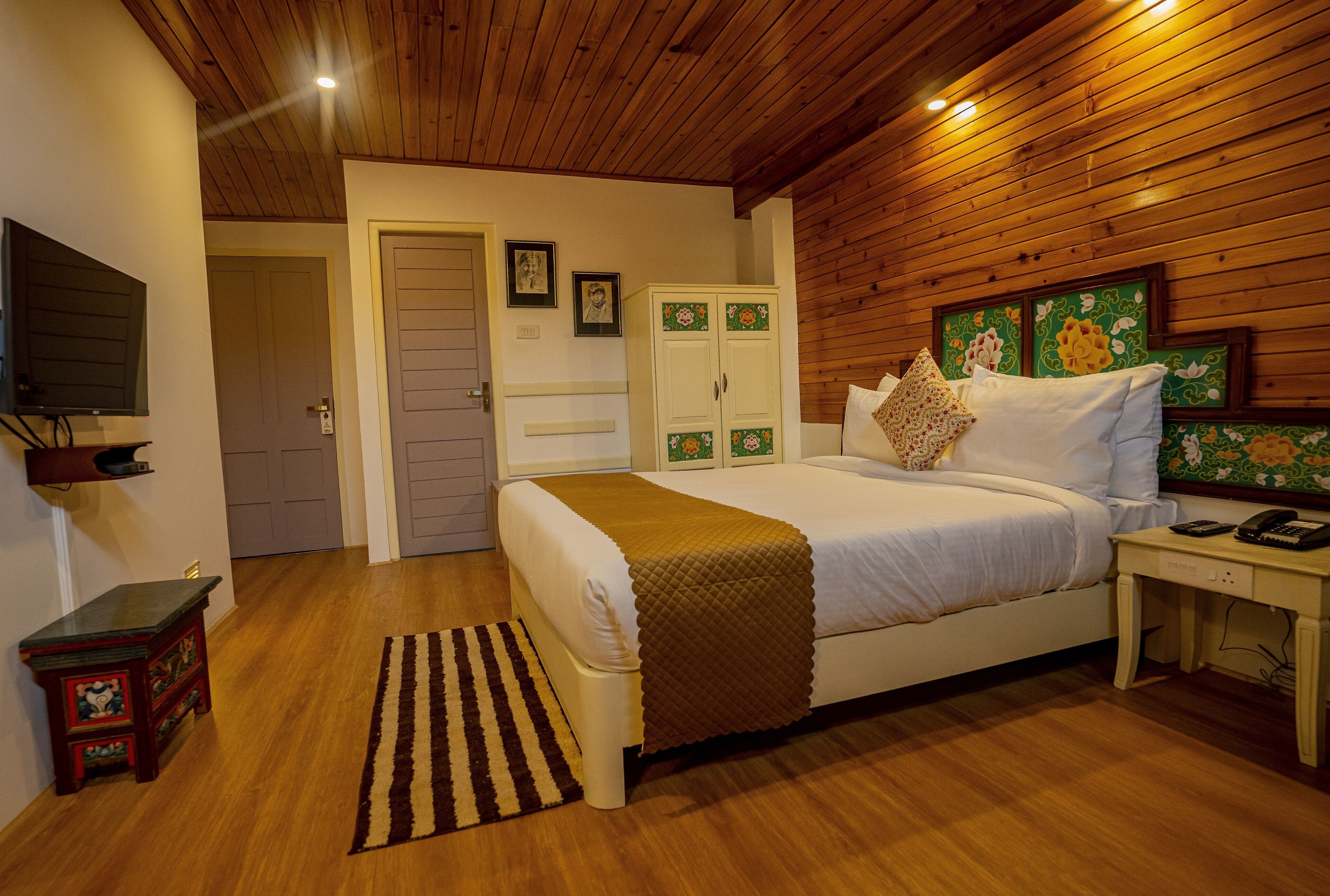 A bedroom with a wooden ceiling, a bed with white bedding, a bedside table with a telephone, and a small cabinet with decorative tiles.