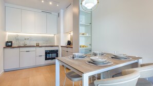 A modern kitchen with white cabinets, a built-in oven, and a dining area with a wooden table and chairs. - Apartamenty Sun & Snow Plażowa Park (Dziwnów)