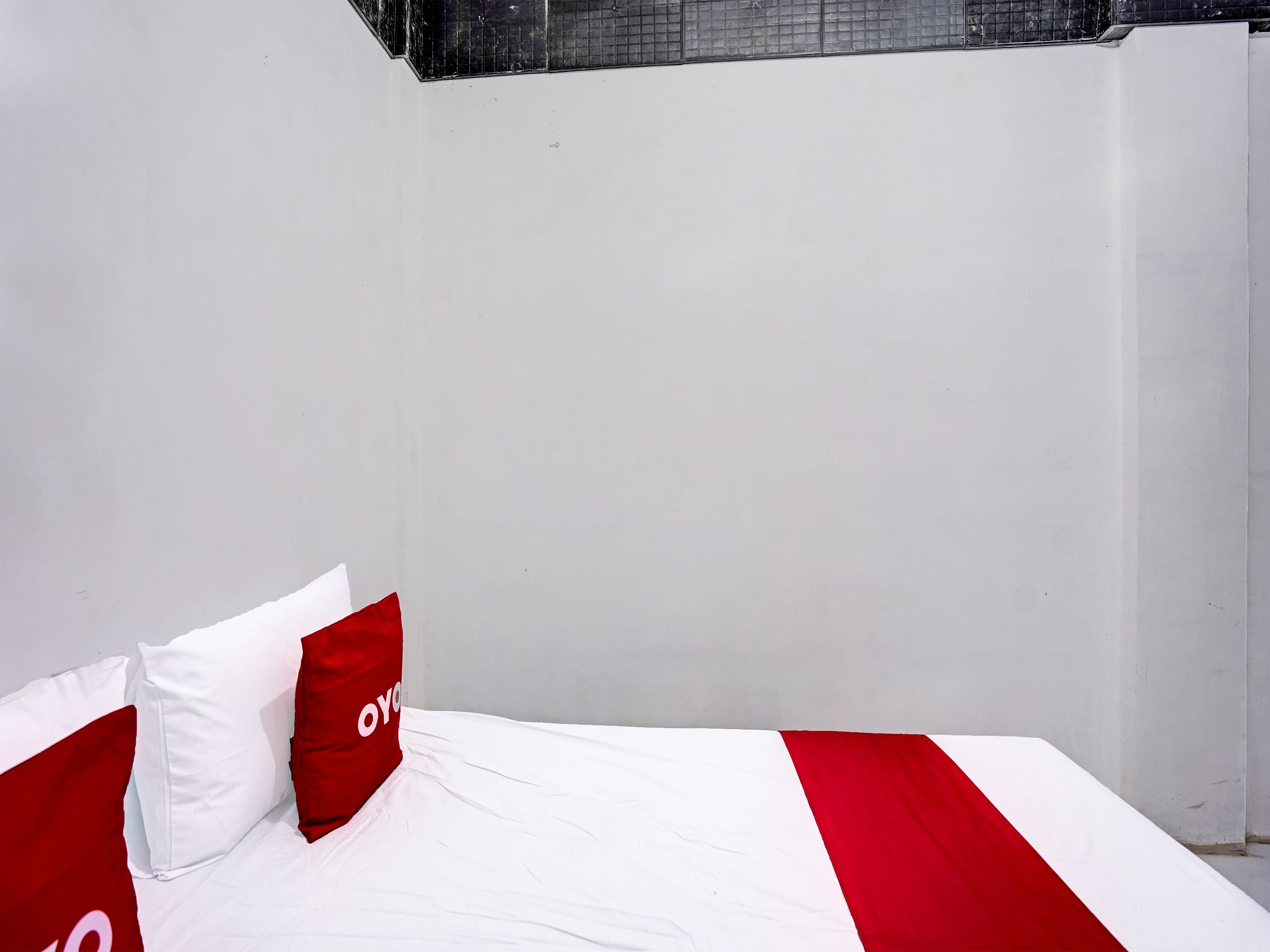 Una cama con colcha y almohadas a rayas blancas y rojas, una pared blanca y una ventana con diseño de cuadrícula.