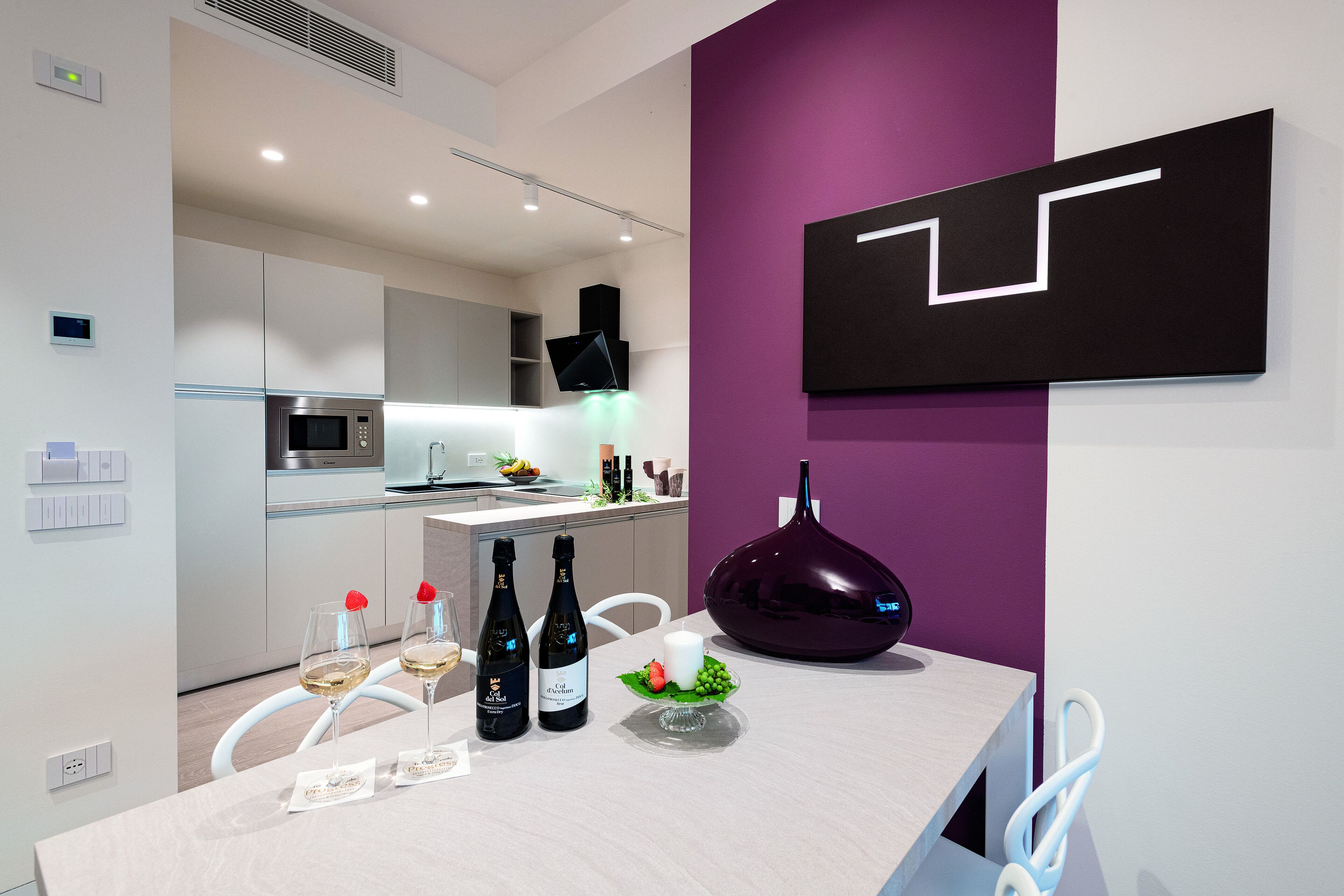 Une cuisine moderne avec un coin repas, agrémentée d’un mur d’accent violet, d’une œuvre d’art géométrique noire et d’une table mise avec des bouteilles de vin et une bougie.