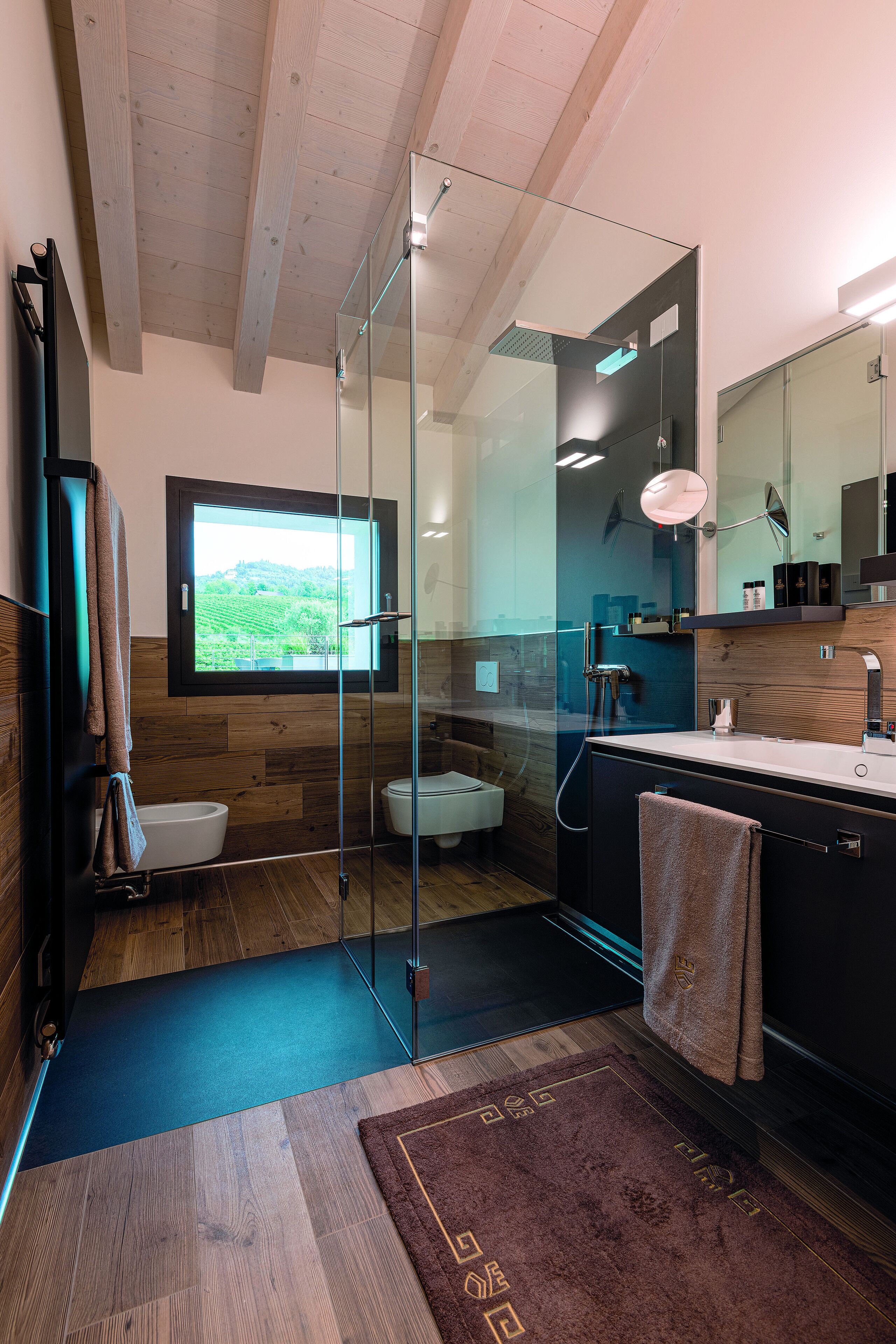 Un baño moderno con cabina de ducha de vidrio, un espejo grande y vistas a un paisaje verde a través de una ventana.