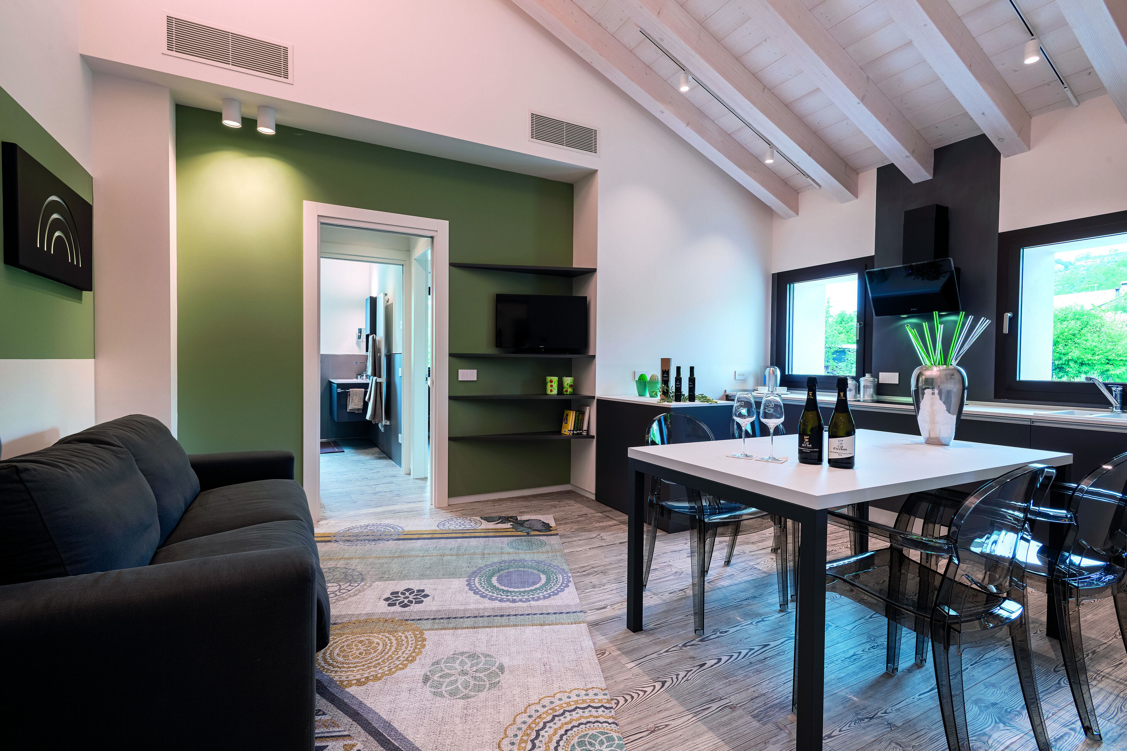 A modern living room with a black sofa, a dining table with chairs, a TV mounted on the wall, and a kitchen area with a window.