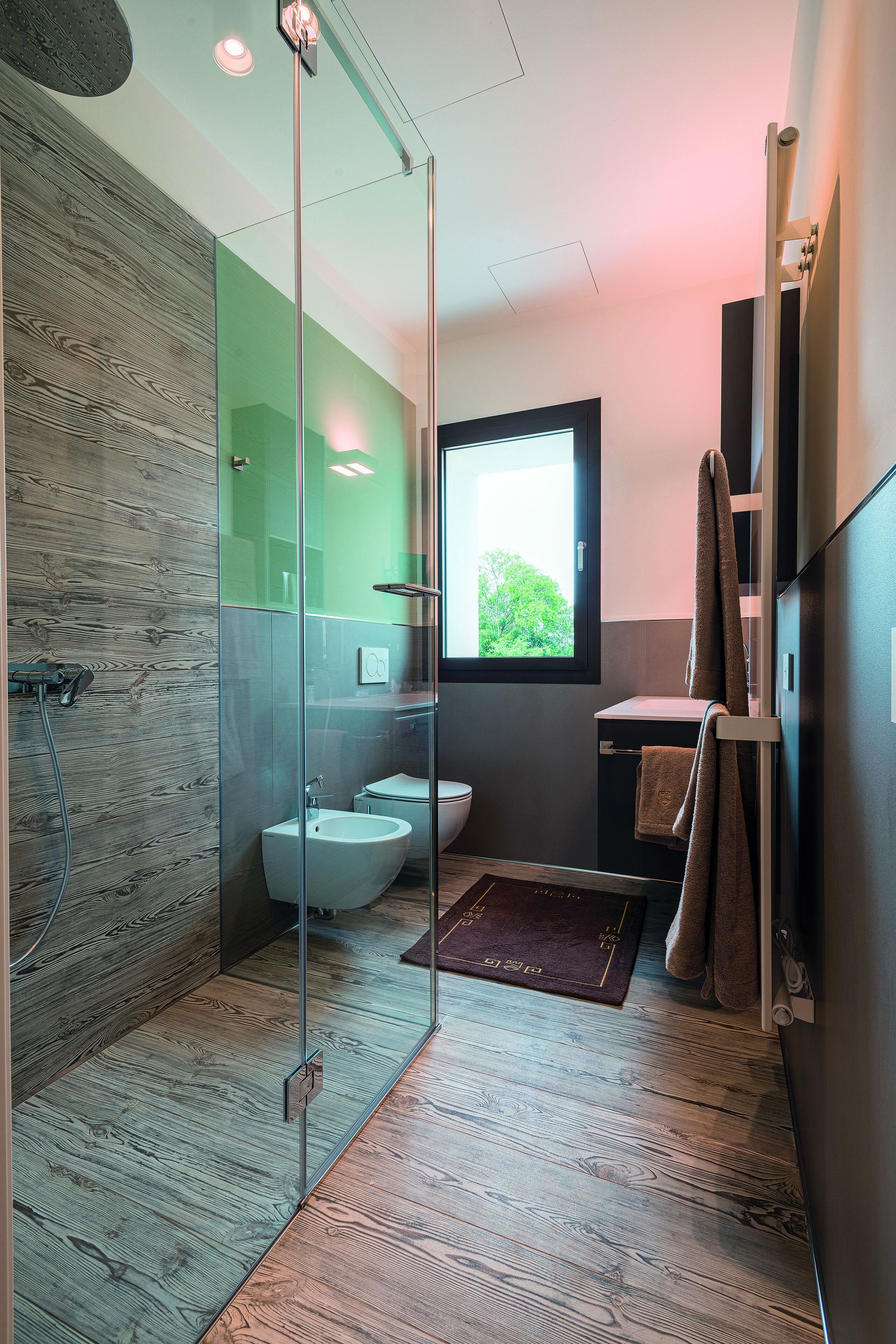 A modern bathroom with a glass shower enclosure, a bidet, and a window providing natural light.