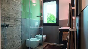 A modern bathroom with a glass shower enclosure, a bidet, and a window providing natural light.