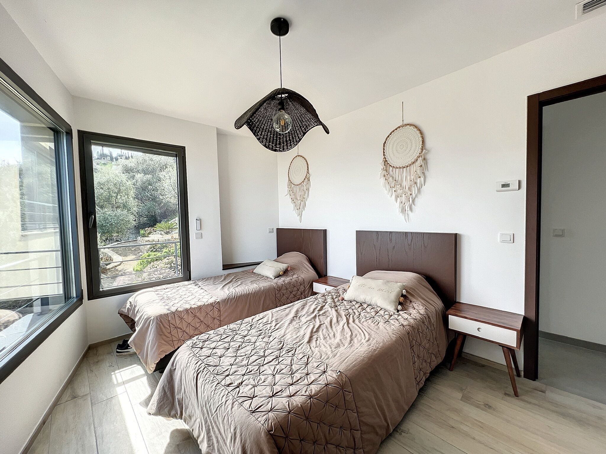 A modern bedroom with two beds, a large window, and a view of greenery.