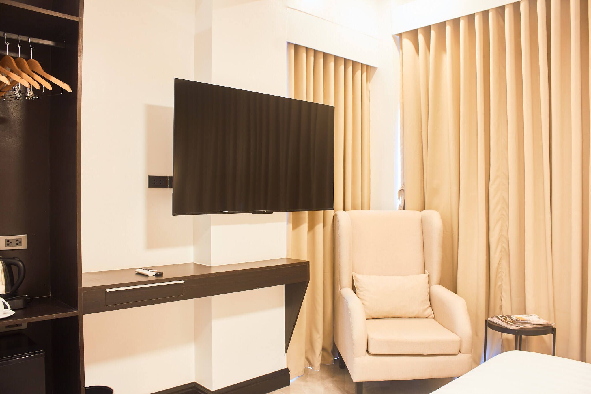 A hotel room with a flat-screen TV mounted on a dark wooden console, a beige armchair, a bed, and beige curtains.