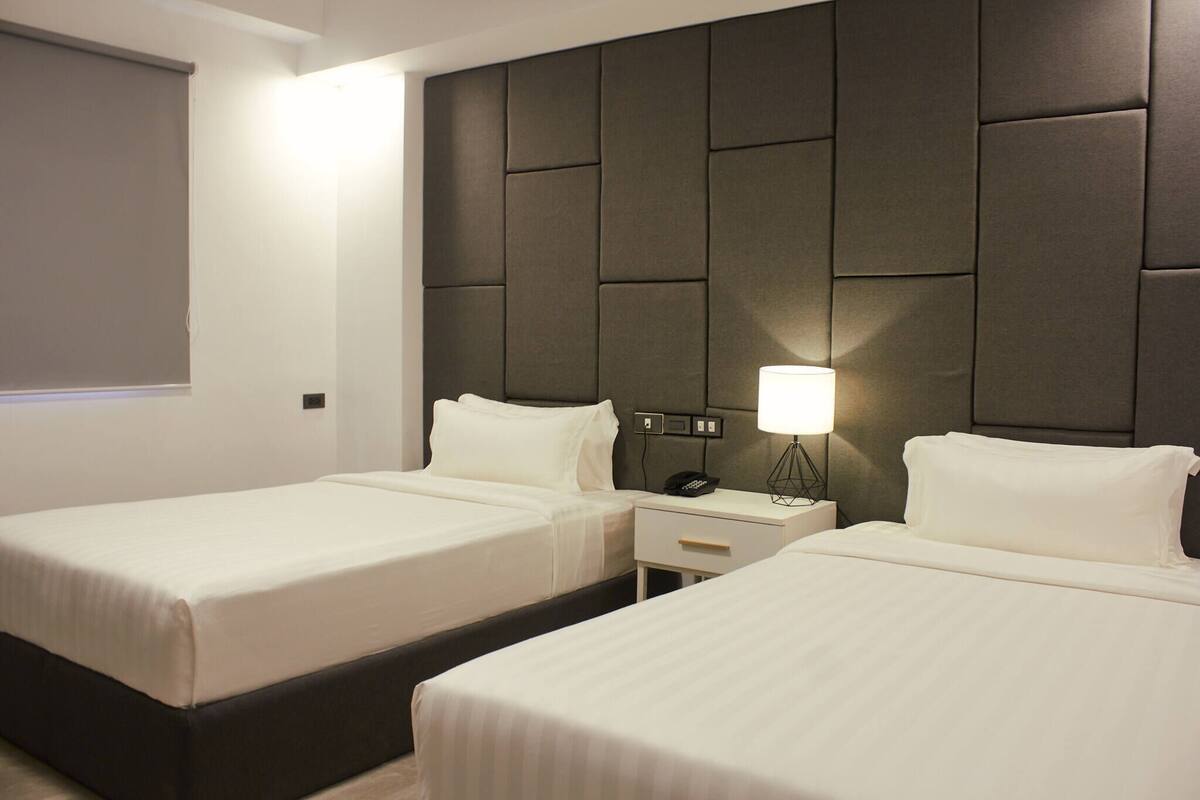 A hotel room with two beds, a headboard with a padded design, bedside tables with lamps, and a window with a blind.