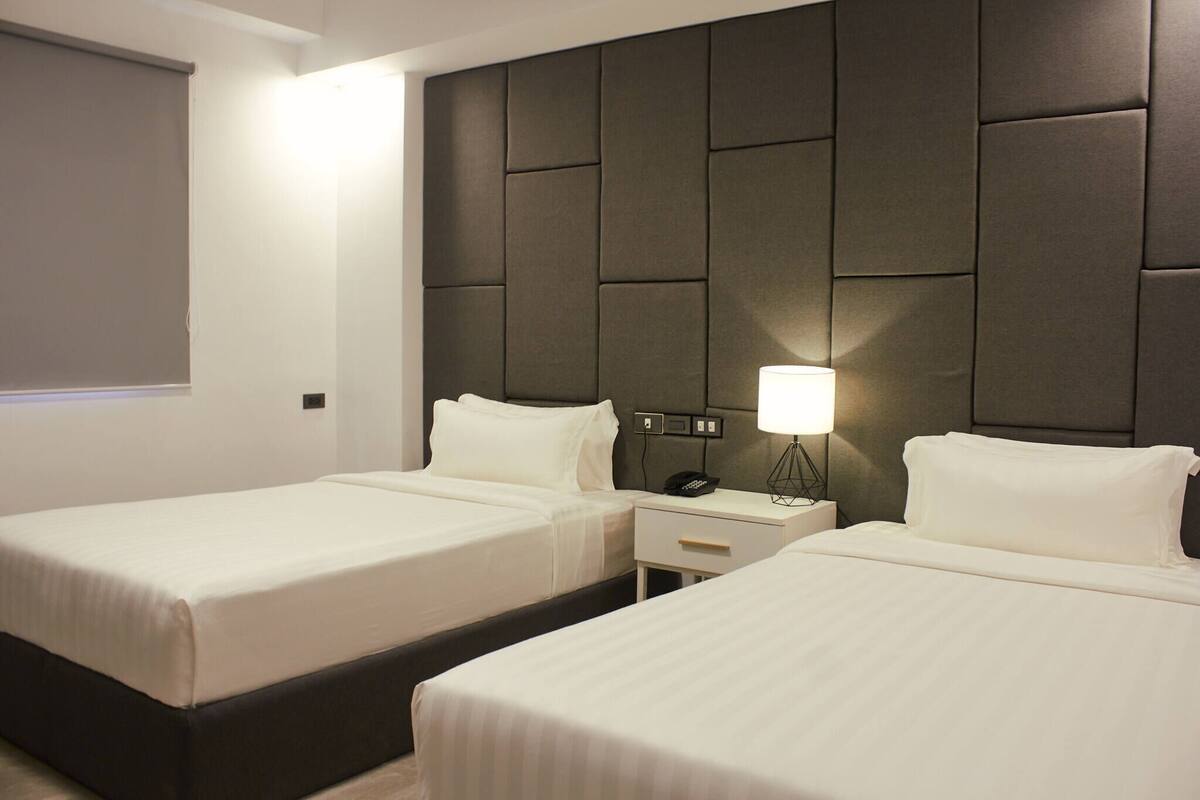 A hotel room with two beds, a headboard, bedside tables, a lamp, and a window with blinds.