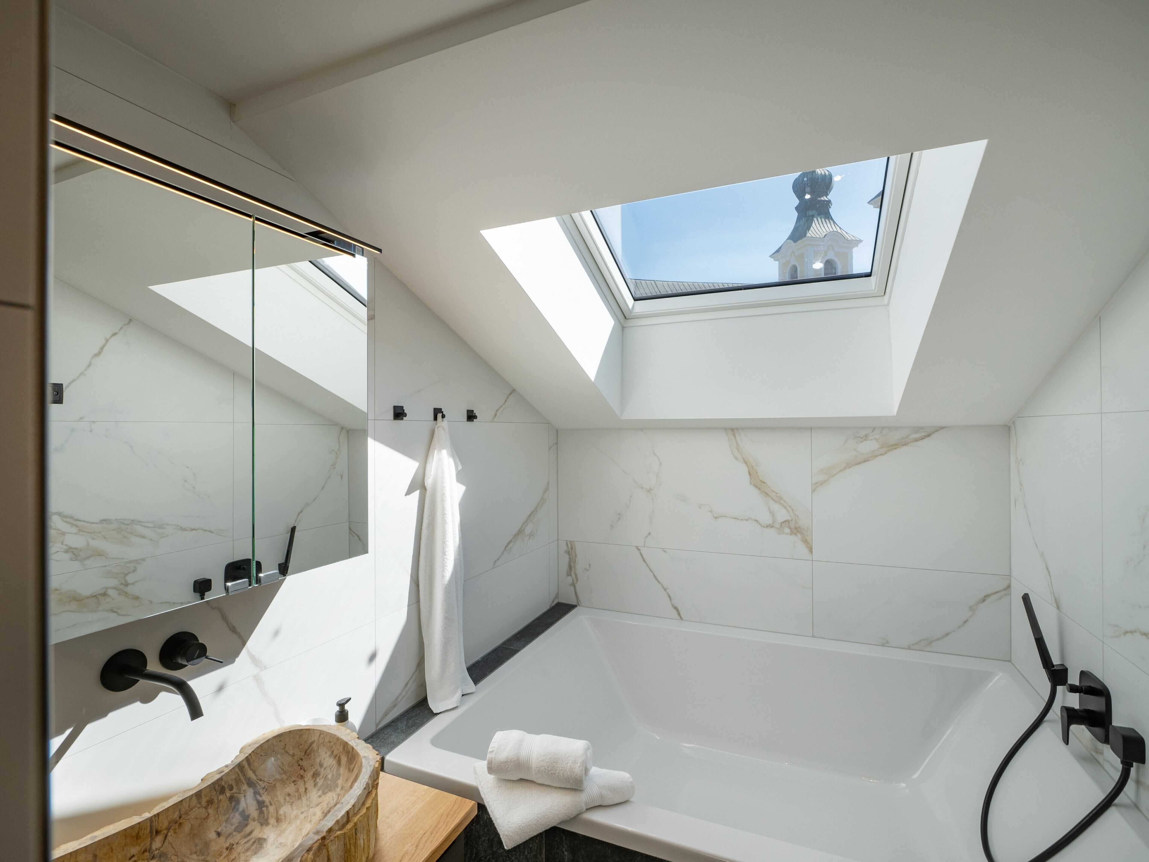 A modern bathroom with a skylight, a bathtub, a vanity with a wooden bowl sink, and marble tiles.