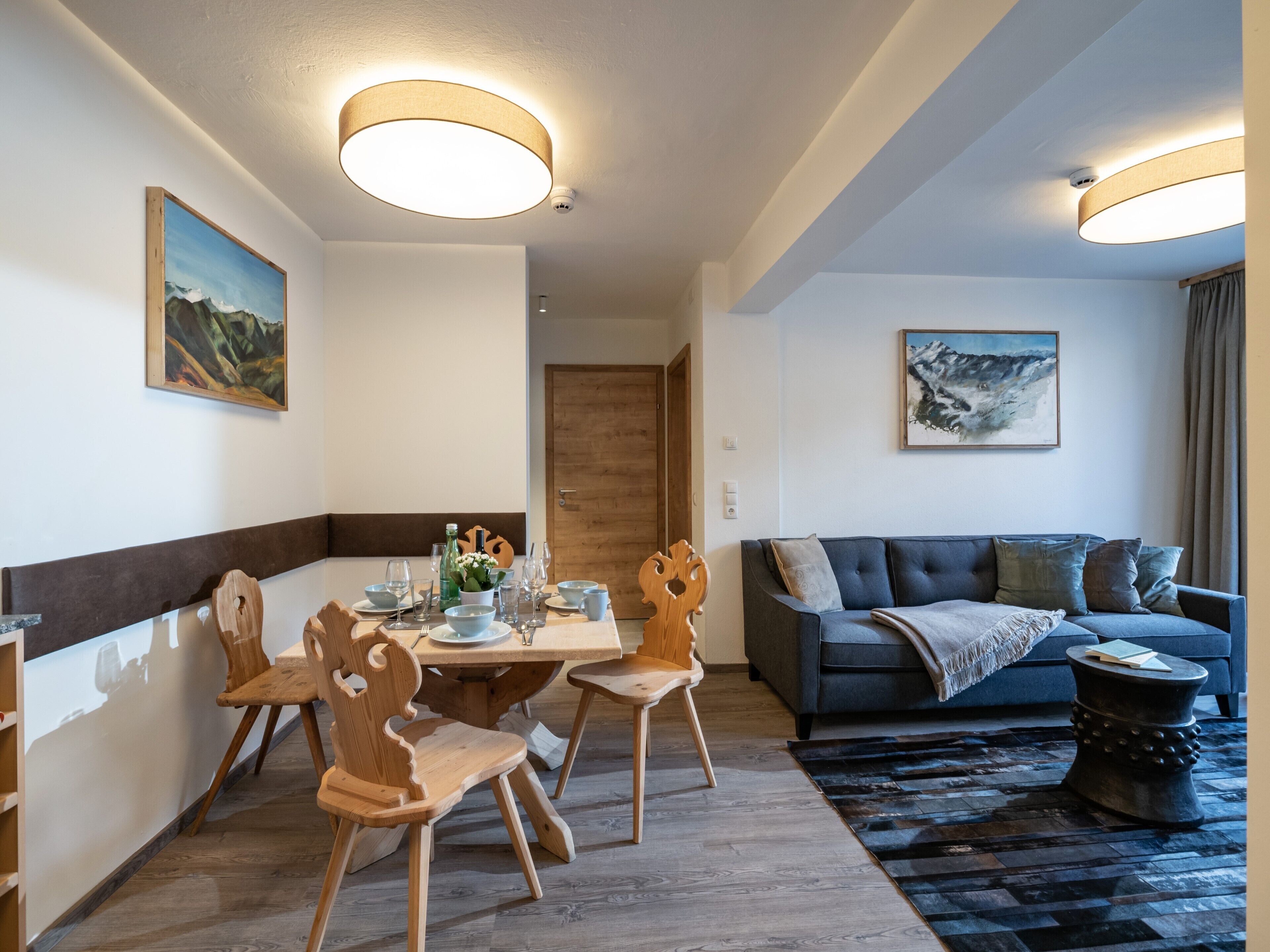 Una sala de estar moderna con un sofá azul oscuro, sillas de madera, una mesa de comedor con platos y vasos, y dos lámparas de techo.