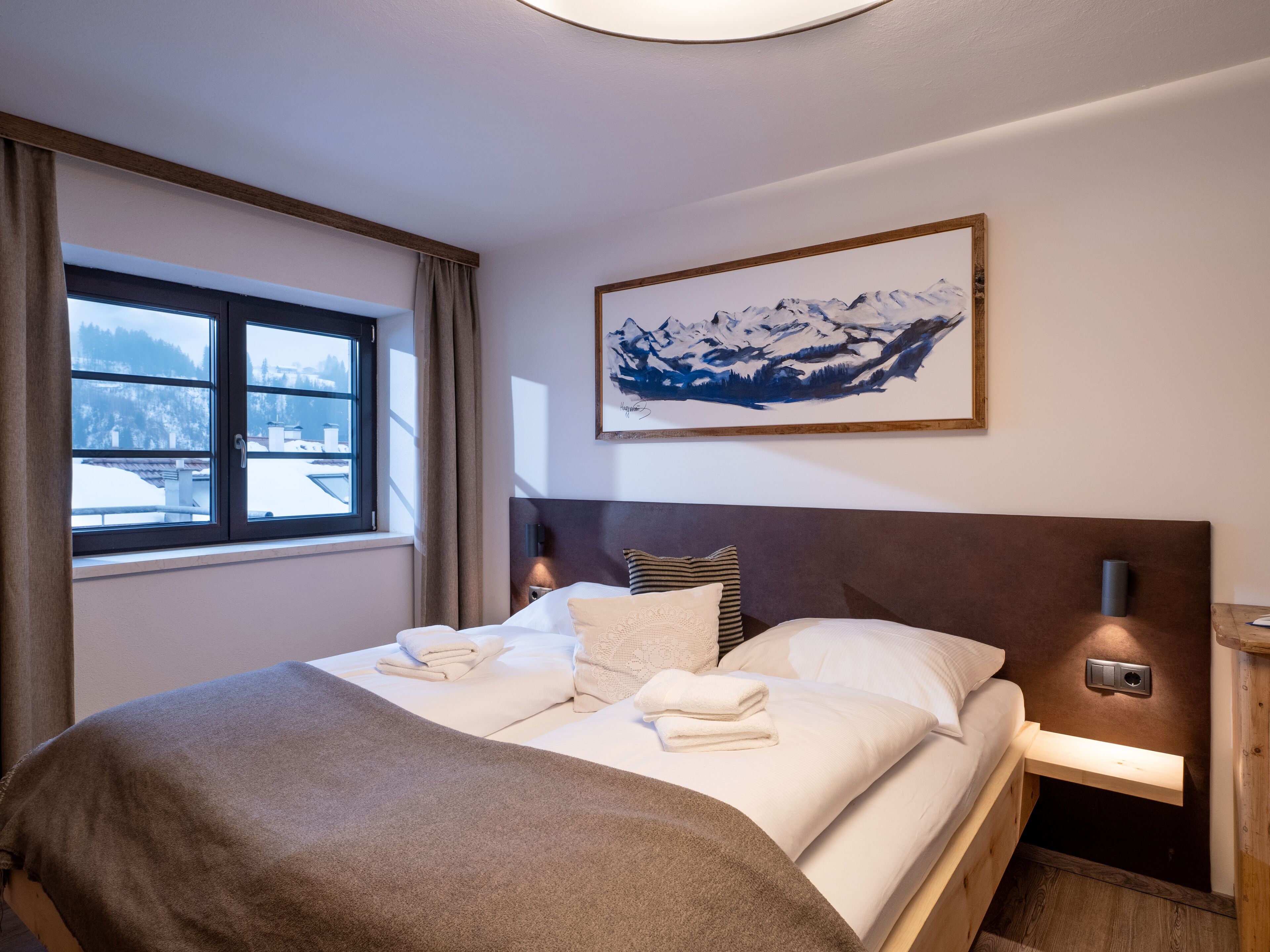 A neatly made bed with towels, a painting of mountains on the wall, and a window with a view of buildings.