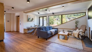 A spacious living area with a sofa, a dining table, and a chair. There is a large window offering a view of a house and trees. - Alpine Homes - Hopfgarten Zentrum (Hopfgarten im Brixental)