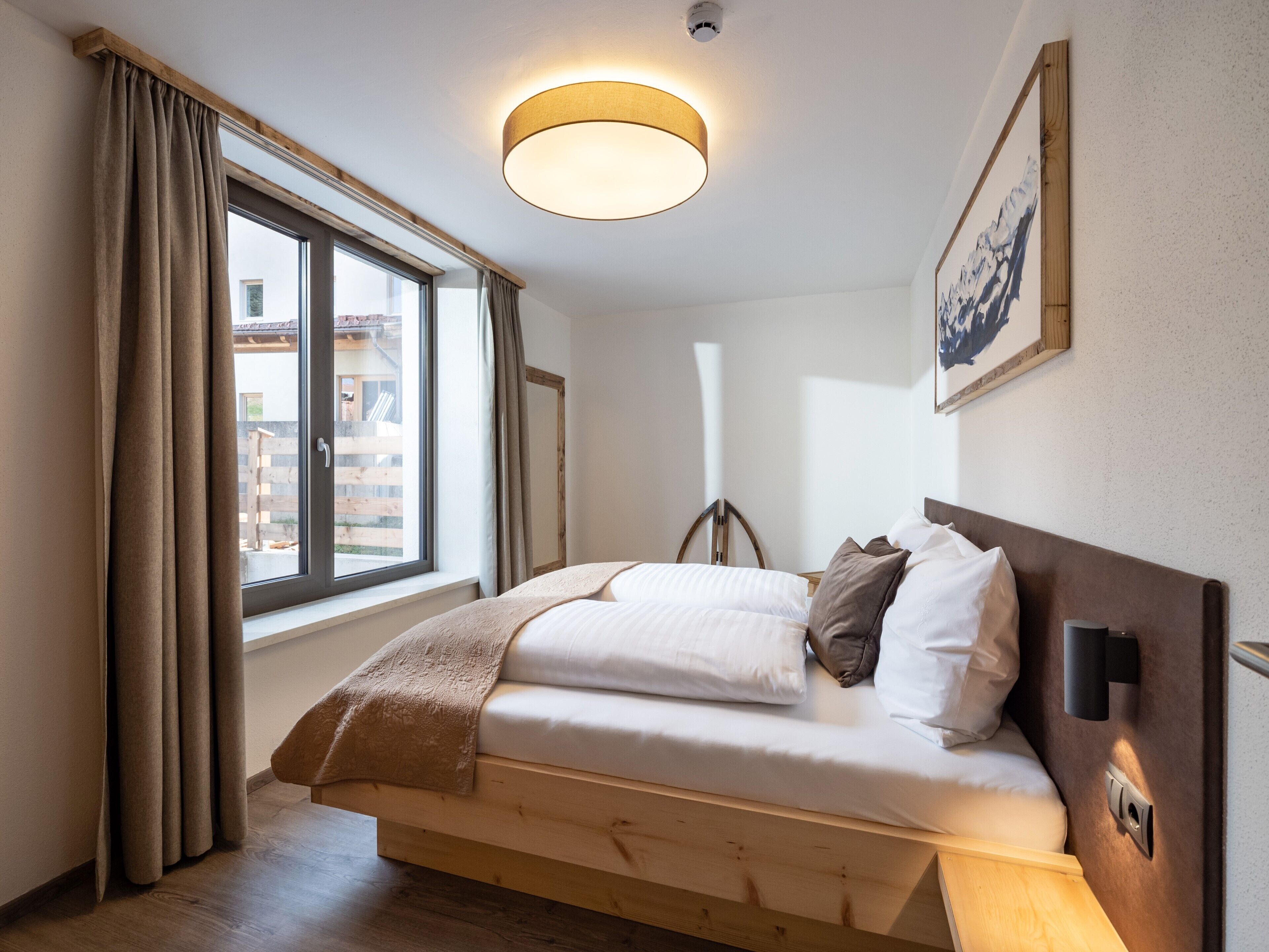 Un dormitorio con una cama de madera, una ventana con cortinas, una lámpara en el techo y un cuadro enmarcado en la pared.