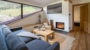 A modern living room with a sofa, a coffee table, a fireplace, and a large window overlooking a green landscape. - Alpine Homes - Hopfgarten Zentrum (Hopfgarten im Brixental)