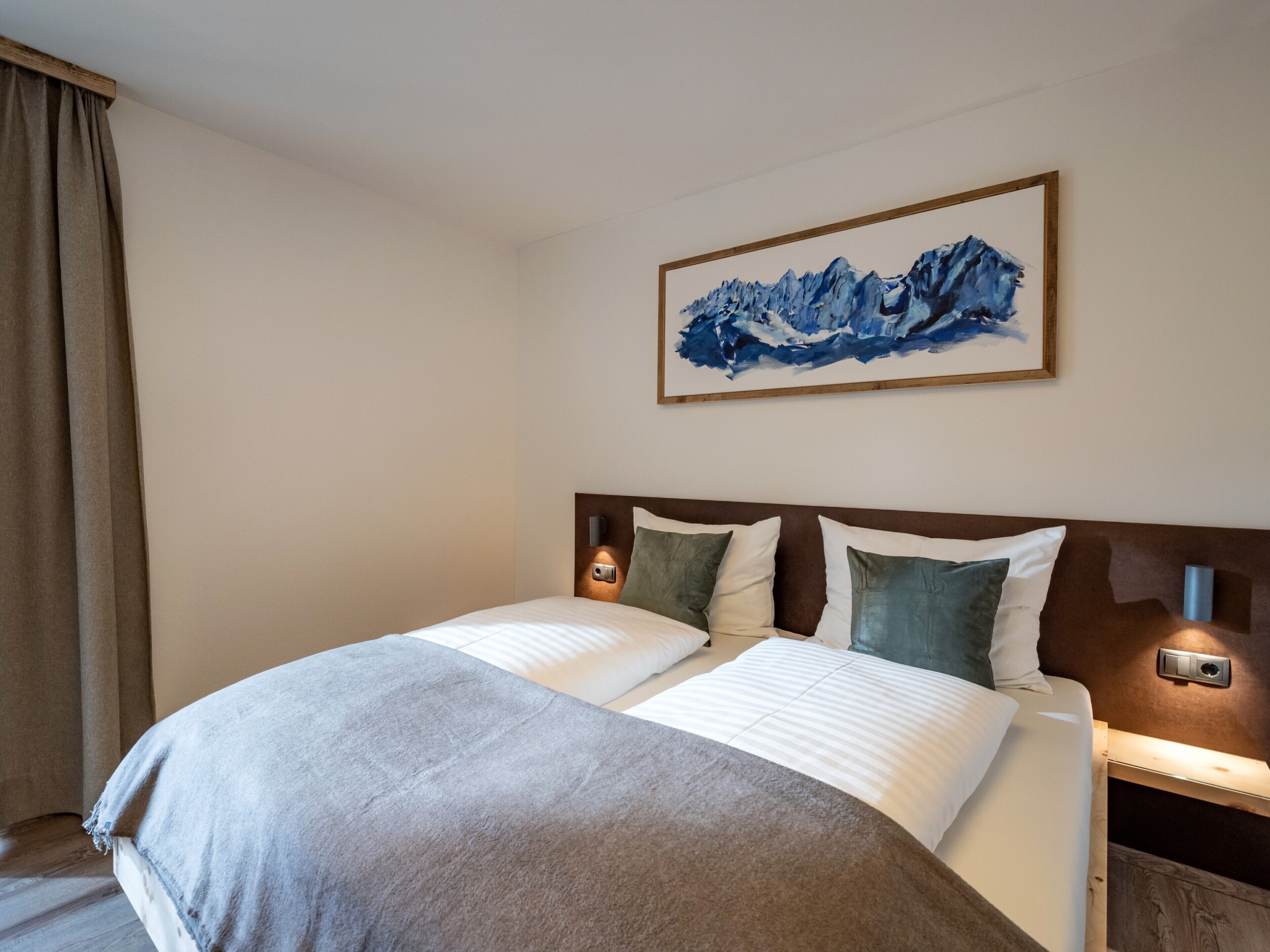 A hotel room with a large bed, a headboard with integrated lighting, a painting of mountains on the wall, and a window with curtains.