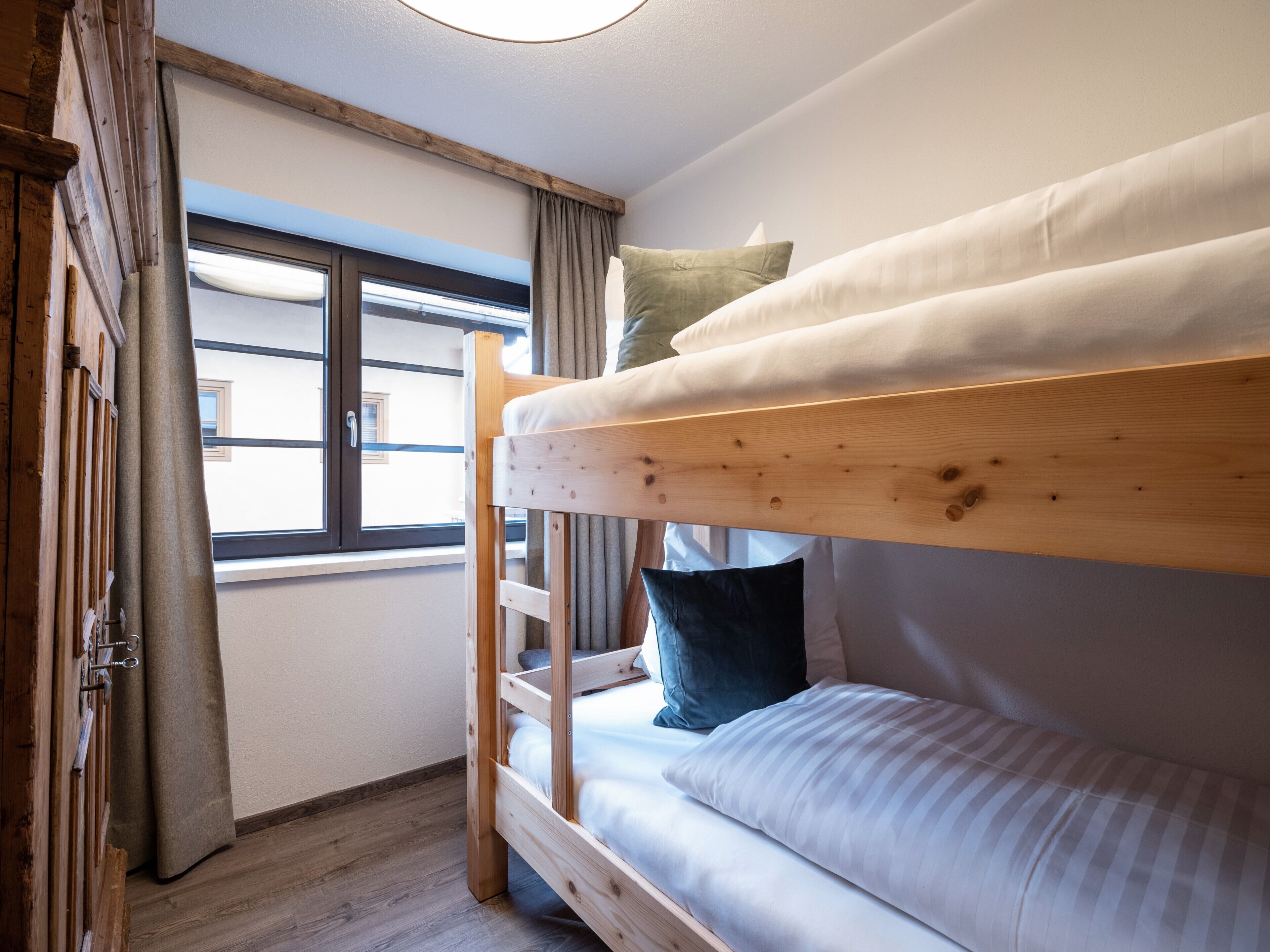 A bunk bed with a wooden frame, a window with curtains, and a view of a building outside.