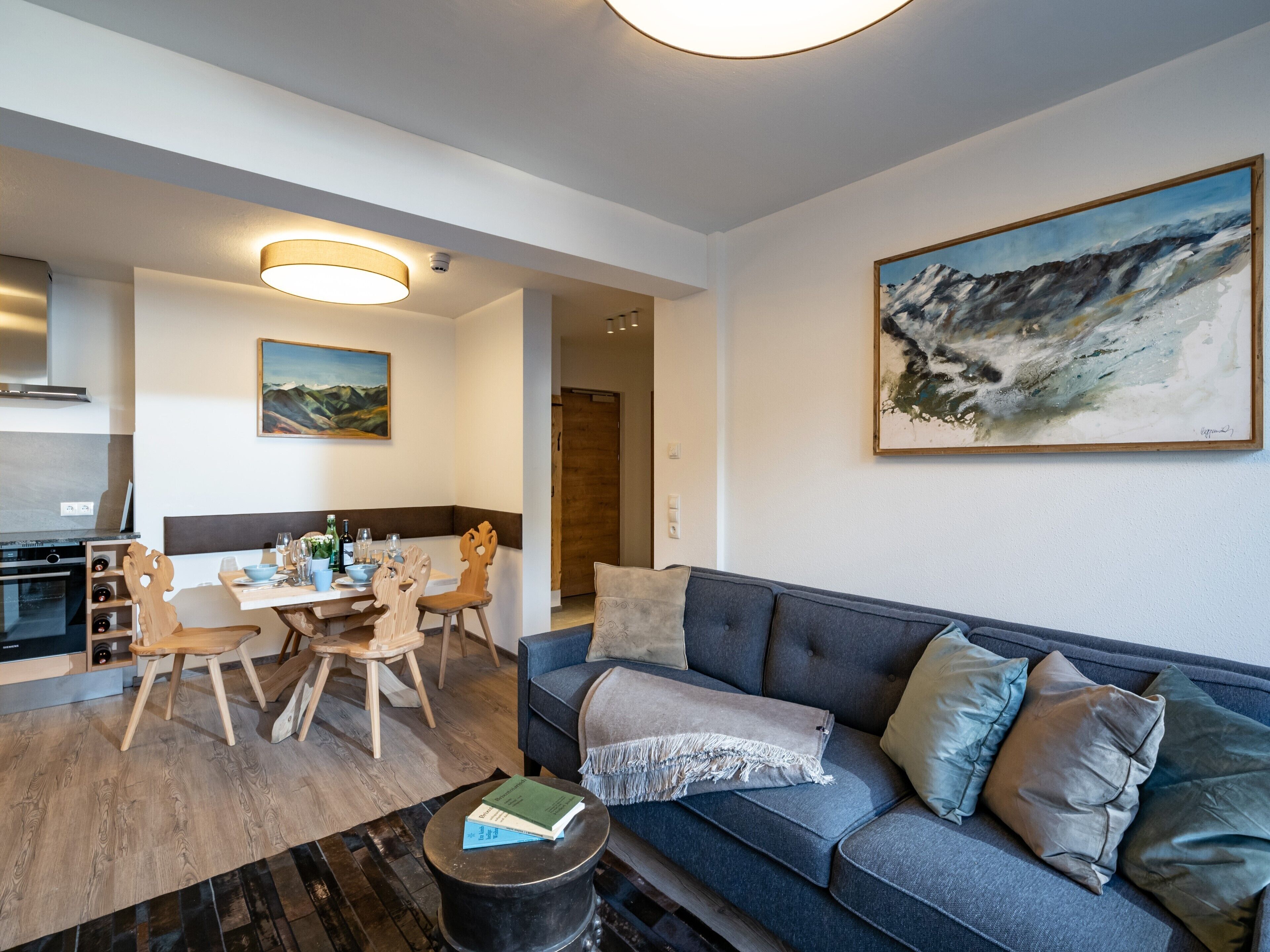 A modern living room with a grey sofa, wooden dining table, and chairs. A painting of a mountain landscape hangs on the wall.