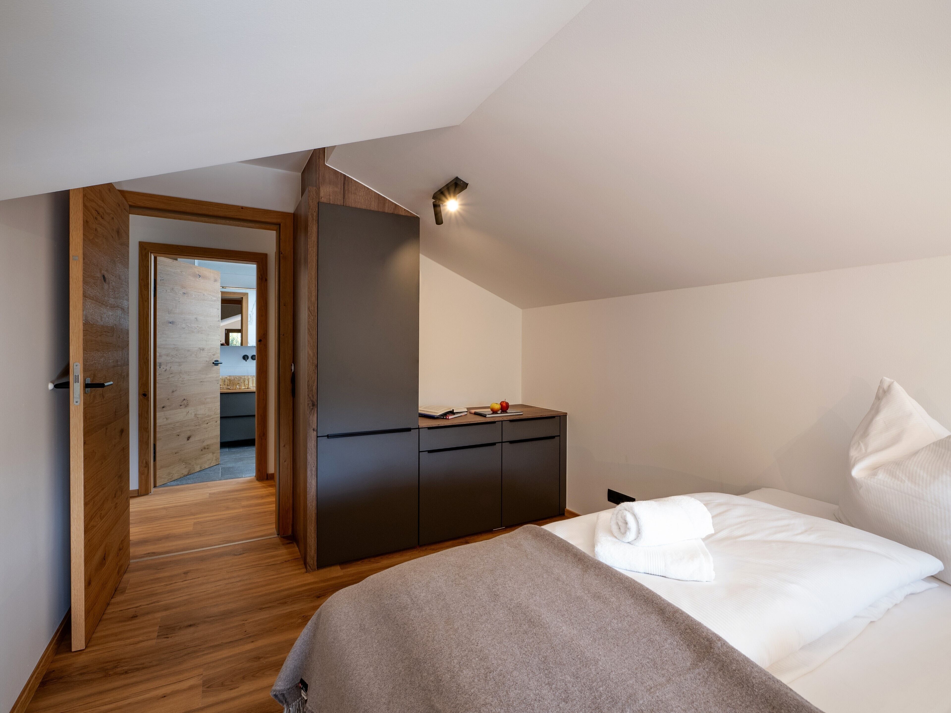 Un dormitorio moderno con suelo de madera, una cama con ropa blanca, un armario oscuro y un escritorio pequeño con un ordenador portátil y fruta.